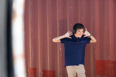 Vergrößerte Ansicht von Ein Junge steht vor einer braunroten Wand, trägt Kopfhörer und hat die Hände an den Kopfhörern.