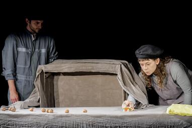 Vergrößerte Ansicht von Eine Bühnenszene: Ein Mann steht links und eine Frau rechts hinter einem Tisch, auf dem ein Puppenhaus aufgebaut ist.