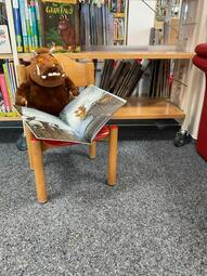 Vergrößerte Ansicht von Der Grüffelo, ein großes bärenartiges Stofftier sitzt auf einem Stuhl und hat ein aufgeschlagenes Buch auf dem Schoß.