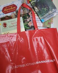 Vergrößerte Ansicht von Eine rote Tasche mit der Beschriftung &quot;Stadtbibliothek Mannheim&quot;. Sie liegt auf einem Tisch und es schauen Bücher für Senioren aus ihr heraus.