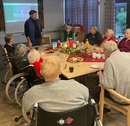 Vergrößerte Ansicht von Senioren sitzen an einem Tisch. Ein Mann steht vor einer Leinwand.