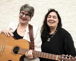Vergrößerte Ansicht von Zwei Frauen, die lachen. Eine von ihnen hält eine Gitarre in der Hand.