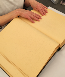 Vergrößerte Ansicht von Ein großes Tastbuch liegt ausgebreitet auf einem Tisch, zwei Hände lesen es.