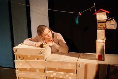 Vergrößerte Ansicht von Auf einer kleinen Bühne aus Holzkisten lässt eine Schauspielerin einen Plüschhund zu einem Leckerli hinaufblicken. Das Leckerlin liegt oben in einem Haus aus aufgetürmten Pappboxen.