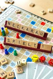 Vergrößerte Ansicht von Auf einem Scrabble-Spielbrett Spielfiguren, Würfel, Spielkarten und Srabble-Plättchen, die die Worte "Neckarau spielt!" bilden.