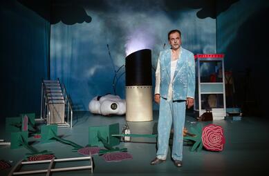 Vergrößerte Ansicht von Eine Person in einem blau-weißen Anzug im Wolkenmuster steht auf der Bühne. Um ihn herum liegen umgefallene 2D-Rosen und ein Teil eines Schiffsgeländers. Ein Wolkenauto, eine Telefonzelle und eine umgefallene Leiter sind im Hintergrund zu sehen,…