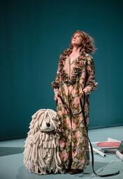 Vergrößerte Ansicht von Eine Frau in einem eleganten Gewand mit Blumenmuster. Sie hat eine Puppe eines weißen, zotteligen Hundes an ihrer Seite. Ihre Miene ist arrogant, Kinn leicht gehoben.Im Hintergrund sind 2D-Rosen auf dem Boden verstreut zu sehen.