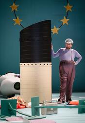 Vergrößerte Ansicht von Eine Person in einem fliederfarbenen Hemd und plaumenfarbener Hose lehnt gegen ein Rohr, das einem Auspuffrohr eines Frachters ähnelt. Der Mann trägt eine silberne Kapitänsmütze und schaut verblüfft in die Ferne. Der Hintergrund ist hellblau mit im…