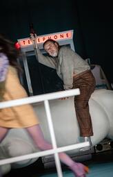 Vergrößerte Ansicht von Ein Mann in einem grauen Pullover, brauner Hose und silbernen Stiefeln sitzt auf einer weißen Wolke. In seiner rechten Hand hält er einen Gegenstand hoch, der im dunklen Hintergrund verschwindet. Im Hintergrund ist das beleuchtete Schild einer…