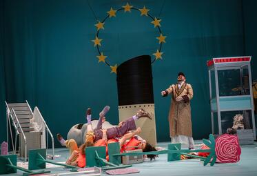 Vergrößerte Ansicht von Das Bühnenbild zeigt das Symbol der EU auf hellblauen Hintergrund. Auf der Bühne steht ein Auspuffrohr eines Schiffes, eine umgefallene Leiter und sechs umgefallene 2D-Blumenaufsteller. Eine Gruppe von drei Personen liegt in Rettungswesten verknotet…