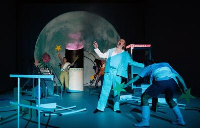 Vergrößerte Ansicht von Ein Mann in einem blau-weißen Anzug öffnet seinen Mund in einem dramatischen Schrei. Das Bühnenbild zeigt einen großen Vollmond im Hintergrund. Ein Auspuffrohr eines Schiffes steht neben einer umgefallenen Leiter, einer Telefonzelle und zwischen…