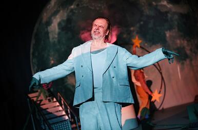 Vergrößerte Ansicht von Dramatisches magentafarbenes Licht scheint einem Mann in einem blau-weißen Anzug in das Gesicht.Er kneift schmerzverzogen die Augen zusammen, Arme in einer eleganten Bewegung gehoben. Im Hintergrund sieht man einen großen Vollmond.