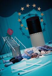 Vergrößerte Ansicht von Das Bühnenbild zeigt einen goldenen EU-Stern auf hellblauen Hintergrund. Das Auspuffrohr eines Schiffes steht neben einer umgefallenen Leiter. Ein 2D-Blumenaufsteller und ein umgefallenes Stück Geländer liegen verstreut herum. Eine Gruppe an…