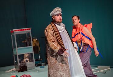 Vergrößerte Ansicht von Eine Person in einem eleganten, braunen Mantel und einer silbernen Kapitänsmütze schaut in Richtung des Betrachters. Eine zweite Person läuft hinter ihm und zieht eine Grimasse. Die zweite Person trägt eine orangene Rettungsweste.