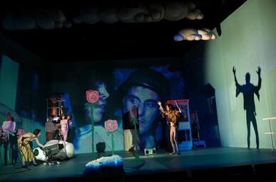 Vergrößerte Ansicht von Das Bühnenbild zeigt einen dunklen Raum mit blauen Wänden. 2D-Rosenaufsteller sind zu sehen, sowie eine Telefonzelle und eine Leiter. Eine Person steht auf  der Bühne vor einem Scheinwerfer, sein Schatten wirft eine scharfe Silhouette auf die rechte…