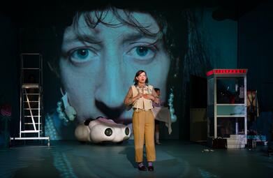 Vergrößerte Ansicht von Eine Person in einem braun-beigen Anzug hält die Hände in Schock gegen die Brust und der Schock spiegelt sich ebenfalls in ihrem Gesicht wider.Auf der Wand hinter ihr wird das Gesicht von einem Mann projiziert.