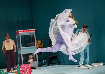 Vergrößerte Ansicht von Eine Person in einem pastell-lila Bodysuit, der unter  einem weißen Kleid getragen wird, springt in die Luft. Das weiße Kleid flattert dramatisch. Zwei weitere Personen stehen in Hintergrund, eine Person schaut die Person im Kleid verwirrt an. Die…