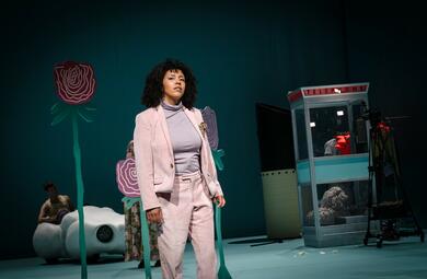 Vergrößerte Ansicht von Eine Person in einem rosa Anzug blickt mit ernster Miene in die Ferne. Das Bühnenbild besteht aus hellblauen Wänden. Eine Telefonzelle ist steht im Hintergrund auf der linken Seite vom Betrachter ausgehend, sowie ein Wolkenauto und 2D-…