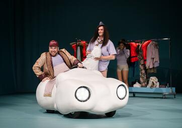 Vergrößerte Ansicht von Ein Mann in einem eleganten braunen Mantel und einer dunkelroten Schildkappe sitzt in einem Wolkenauto. Eine Person mit langen, braunen Haaren,violetter Bluse und Krawatte, der keine Hose trägt, steht grinsend hinter ihm. Im Hintergrund steht eine…