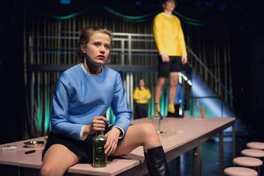 Vergrößerte Ansicht von Camille Dombrowsky (Nora) sitzt mit ernstem Gesichtsausdruck auf einem Tisch, Krakowski, Zastrau und Brück stehen im Hintergrund.