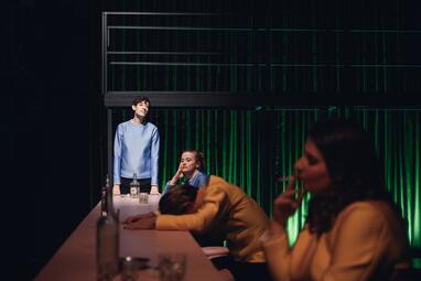 Vergrößerte Ansicht von Camille Dombrowsky (Nora), Robin Krakowski (Tom) und Sarah Zastrau (Vera) sitzen gelangweilt an einem Tisch, Rocco Brück (Helge) steht am Tischende.