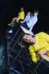 Vergrößerte Ansicht von Robin Krakowski (Tom) liegt auf einem Tisch, Sarah Zastrau (Vera), Camille Dombrowsky (Nora) und Rocco Brück (Helge) sitzen im Hintergrund.