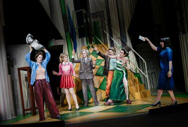 Vergrößerte Ansicht von Eine entblößte Frau hält drohend eine Statue über ihren Kopf. Vier andere Frauen gestikulieren ihr warnend und entsetzt zu. Eine Frau im blauen Kleid redet auf sie ein, mit Handy in der Hand..