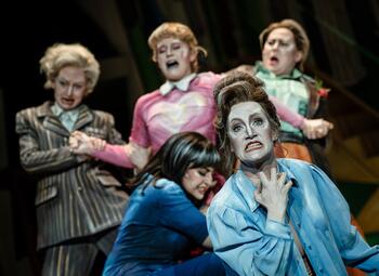 Vergrößerte Ansicht von Frau in blauer Bluse aufgelöst und wütend im Vordergrund. Drei andere Frauen halten eine kreischende Vierte im Hintergrund zurück.