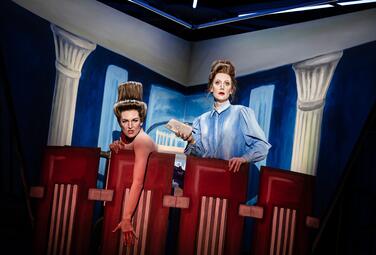 Vergrößerte Ansicht von Zwei Frauen blicken über ein Geländer. Eine Frau hat eine seltsame Frisur in der Form eines Hutes. Die andere trägt eine blaue Bluse und hält ein Handy in der Hand.