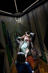 Vergrößerte Ansicht von Frau in Lederjacke streckt die Arme in einer dramatischen Pose über ihren Kopf, während sie von einer Frau in einem blauen Kleid sinnlich am Körper berührt wird.