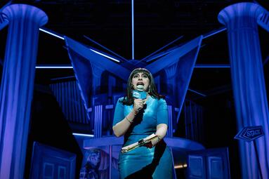 Vergrößerte Ansicht von Frau in einem blauen Kleid hält eine Ansprache in ein Mikrophon. Der Hintergrund ist in blaues Licht getaucht.