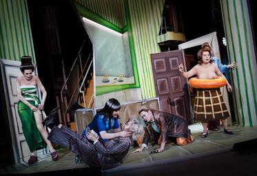 Vergrößerte Ansicht von Frau in einem blauen Kleid würgt Frau in einem grauen Anzug. Frau in einem grünen Kleid und Frau in Lederjacke schauen zu. Eine weitere Frau steht Oberkörper frei mit einem Schwimmring um die Hüfte neben dran und starrt mit gehobenen Zeigefinger in…