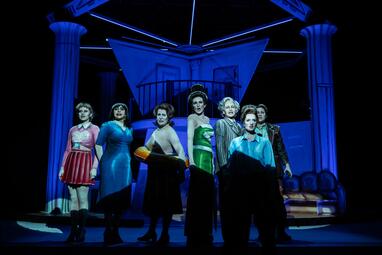 Vergrößerte Ansicht von Dramatisches blaues Licht scheint auf sieben Frauen, die mit ernster Miene in den Zuschauerraum blicken und singen.