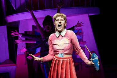 Vergrößerte Ansicht von Frau in einem rosa Cheerleading-Outfit hält einen blauen Slushie. Singend ist ihr Mund weit auf und zeigt eine blau-gefärbte Zunge. Im Hintergrund sieht man eine Schlange an Menschen, die in einer Formation die Hände ausstrecken.