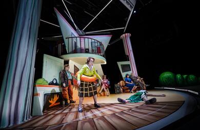 Vergrößerte Ansicht von Das zweistöckige Bühnenbild zweigt unten einen Konferenzraum und oben Geländer. Details wie Türen, Kamin oder Stühle sind auf die Wände nur aufgemalt. Im Vordergrund steht eine Frau in grüner Bluse und braunem Rock. Sie hat einen Schwimmring um die…