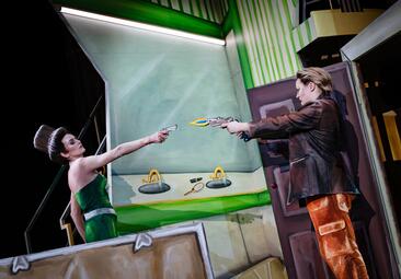 Vergrößerte Ansicht von Eine Frau in grünem Kleid und Haaren, die in die Form eines Hutes frisiert wurden, zielt mit einer Pistole auf eine Frau in Lederjacke und kurzen Haaren, ebenfalls mit Waffe in der Hand. Die Waffen sind zwei-dimensionale, bemalte Plattenausschnitte…