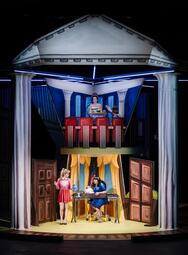 Vergrößerte Ansicht von Das Bühnenbild zeigt zwei Ebenen. Oben sieht man ein Podium, in dem eine Frau in einem grünen Kleid eine Rede hält. Eine Reporterin in blauer Bluse zeichnet ihre Rede auf. Auf der unteren Ebene sieht man ein Büro. Eine Frau in einem blauen Kleid…