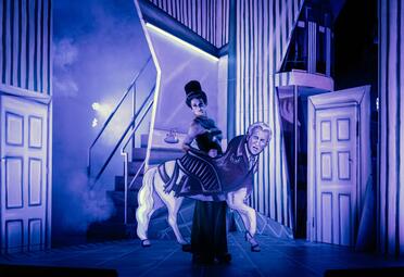 Vergrößerte Ansicht von Eine Frau hat die Haare wie einen Hut gestylt. Sie trägt ein elegantes Kleid und steht hinter einem Pferd, dass den Kopf von George Bush hat. Das Bild ist in dramatisches blaues Licht getaucht.