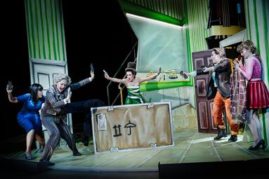 Vergrößerte Ansicht von Ein Tumult in einem WC. Eine Frau in einem grünen Kleid bedroht vier weitere Frauen mit einer Waffe. Die vier Frauen halten panisch, geduckt Abstand. Darunter befindet sich eine Frau in Lederjacke, die einen Bunsenbrenner Richtung Frau in grün…