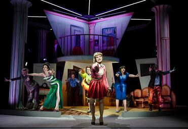 Vergrößerte Ansicht von Sieben Frauen strecken die Zeigefinger in einem choreografischen Gesang aus. Im Vordergrund singt eine Frau in einem rosa Cheerleading-Outfit.