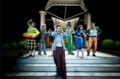 Vergrößerte Ansicht von Eine Frau in blauer, aufgeknöpfter Bluse, hält sich in Entsetzung die Hände an den Kopf. Hinter ihr stehen sechs weitere Frauen. Fünf von denen zeigen ebenfalls auch verschiedene Weisen Entsetzung. Die sechste Frau, in grüner Bluse, braunem Rock und…