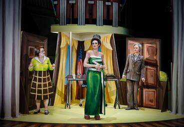 Vergrößerte Ansicht von In einem Office steht eine Frau in einem grünen Kleid und hochgestylten Haaren. Rechts von ihr eine Frau mit kurzen Haaren und einem grauen Anzug, links von ihr eine nervöse Frau in grüner Bluse und braunem Rock. Das Bühnenbild ist zwei-dimensional…