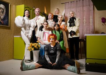 Vergrößerte Ansicht von Neun Schauspieler*innen in bunten Kostümen posieren als Gruppe in einer Küche, die eingerichtet ist, als wäre sie aus den 50er Jahren.