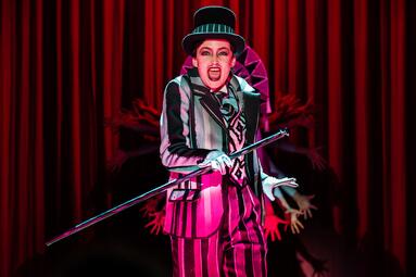 Vergrößerte Ansicht von Annemarie Brüntjen steht auf der Bühne und hält einen eisernen Gehstock in ihrer rechten Hand. Sie trägt ein schwarz-weiß gestreiften Anzug mit schwarzen Zylinder. Sie reißt ihren Mund weit auf und blickt zum Publikum. Hinter ihr befindet sich ein…