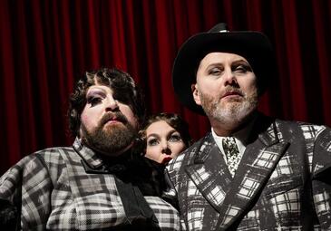 Vergrößerte Ansicht von zwei schick gekleidete Personen schauen zum Publikum. Eine weitere person mit staunender Miene schaut hinter ihnen hervor.