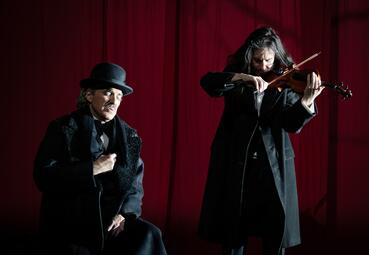 Vergrößerte Ansicht von Ein Mann in einem schwarzen langen Mantel und mit einem Hut hält sich ergriffen die Hand an das Herz. Rechts neben ihm steht ein Mann mit langen schwarzen Haaren und einem schwarzen Mantel, der Geige spielt.