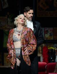 Vergrößerte Ansicht von Man sieht zwei Personen auf der Bühne. Eine Frau in einem silbernen Gewand und mit blonden Haaren, die die Hände in die Hüfte stützt.Hinter ihr steht ein Mann in einem schwarzen  Anzug, der zur rechten Seite schaut.