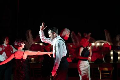 Vergrößerte Ansicht von Ein Mann in einem weißen Hemd steht seitlich zur Kamera. Er streckt einen Arm in die Luft. Hinter ihm sind mehrere Leute zu sehen,von denen ein paar tanzen. Die Bühne ist in rotes Licht getaucht.