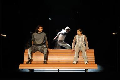 Vergrößerte Ansicht von Zwei Personen in grauen Sportklamotten sitzen auf der Lehne einer Holzbank. Sie schauen sich an. Im Hintergrund rennt eine dritte Person durchs Bild, verschwommen von der Bewegung.