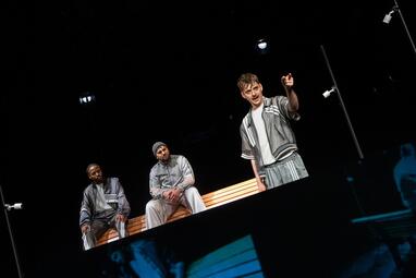 Vergrößerte Ansicht von Drei Personen in grauen Sportklamotten. Zwei Männer sitzen hinten auf der Lehne einer Bank. Der Dritter steht Vorne und deutet mt seinen Zeigefinger in die Ferne.